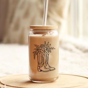 Puede incluir: Un vaso de vidrio transparente con tapa de bamb&uacute; y pajita. El vaso est&aacute; lleno de un l&iacute;quido marr&oacute;n claro y presenta un dibujo lineal negro de una bota de vaquero con detalles florales. El vaso est&aacute; sobre una superficie de madera.