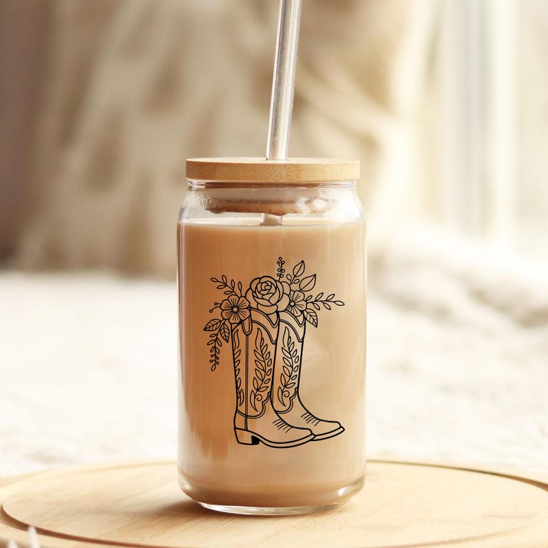 Puede incluir: Un vaso de vidrio transparente con tapa de bamb&uacute; y pajita. El vaso est&aacute; lleno de un l&iacute;quido marr&oacute;n claro y presenta un dibujo lineal negro de una bota de vaquero con detalles florales. El vaso est&aacute; sobre una superficie de madera.