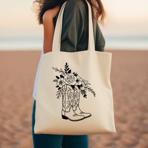 Puede incluir: Bolso tote de color crema con un gr&aacute;fico negro de una bota de vaquero llena de flores. El arreglo floral incluye rosas y otras hojas decorativas. El bolso tiene dos asas y est&aacute; hecho de una tela resistente.
