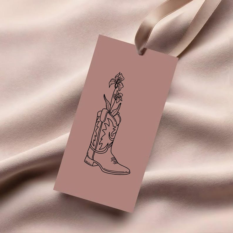 Puede incluir: Una etiqueta rectangular de color rosa polvoriento con un dibujo lineal negro de una bota de vaquero que sostiene flores. La etiqueta est&aacute; suspendida por una cinta de color crema. La bota tiene costuras decorativas.