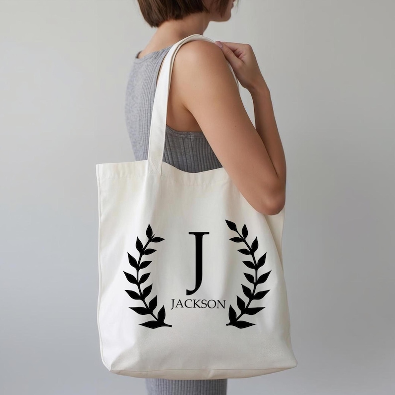 Puede incluir: Bolso tote color crema con una inicial "J" negra y el nombre "Jackson" impreso debajo. La inicial est&aacute; enmarcada por coronas de laurel negras. El bolso se lleva al hombro.