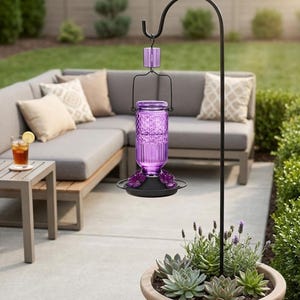 Puede incluir: Un comedero para colibr&iacute;es de vidrio morado con textura de patr&oacute;n de diamantes, que presenta una base de metal negro con puertos de alimentaci&oacute;n en forma de flor, suspendido de un gancho de metal negro anclado en una maceta redonda.