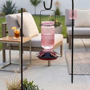 Puede incluir: Un comedero para colibr&iacute;es de vidrio rosa con un dep&oacute;sito de patr&oacute;n de diamantes y una base negra con puertos de alimentaci&oacute;n en forma de flor roja. El comedero cuelga de un gancho de metal negro e incluye un acento de vidrio rosa a juego.