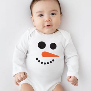 Puede incluir: Un body blanco para beb&eacute; con un dise&ntilde;o de cara de mu&ntilde;eco de nieve. El dise&ntilde;o presenta dos c&iacute;rculos negros para los ojos, un tri&aacute;ngulo naranja para la nariz y una fila de puntos negros para la sonrisa. El body tiene mangas largas.