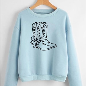 Puede incluir: Sudadera azul claro con un contorno negro de botas de vaquero y un lazo. Las botas tienen costuras decorativas. La sudadera tiene mangas largas y cuello redondo.