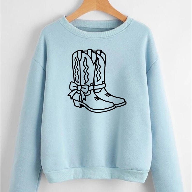 Puede incluir: Sudadera azul claro con un contorno negro de botas de vaquero y un lazo. Las botas tienen costuras decorativas. La sudadera tiene mangas largas y cuello redondo.