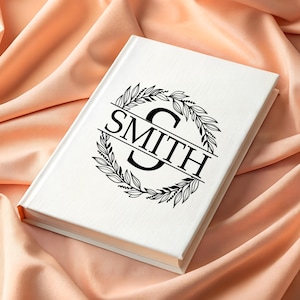 Puede incluir: Un libro blanco con el nombre "SMITH" en letras negras, rodeado por una corona floral negra. El libro descansa sobre una tela color melocot&oacute;n, creando un fondo suave y elegante. El dise&ntilde;o de la portada es de estilo monograma.