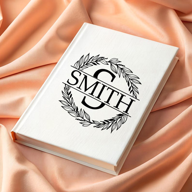 Puede incluir: Un libro blanco con el nombre "SMITH" en letras negras, rodeado por una corona floral negra. El libro descansa sobre una tela color melocot&oacute;n, creando un fondo suave y elegante. El dise&ntilde;o de la portada es de estilo monograma.