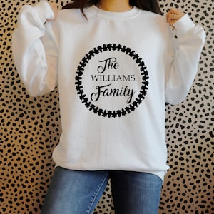 Puede incluir: Sudadera blanca con mangas largas. En la parte delantera, un dise&ntilde;o circular negro con las palabras "The Williams Family" en fuente cursiva, rodeado por un borde negro. El fondo es un estampado de animales en negro y marr&oacute;n.
