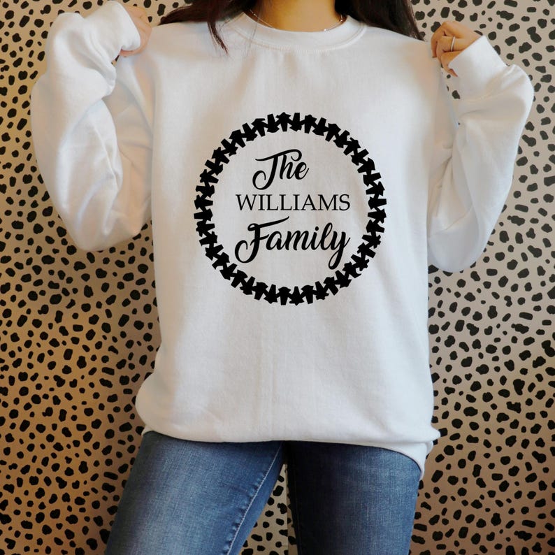 Puede incluir: Sudadera blanca con mangas largas. En la parte delantera, un dise&ntilde;o circular negro con las palabras "The Williams Family" en fuente cursiva, rodeado por un borde negro. El fondo es un estampado de animales en negro y marr&oacute;n.