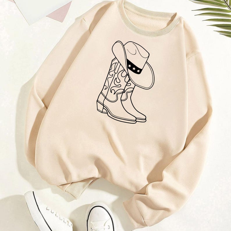 Puede incluir: Sudadera beige con un gr&aacute;fico en blanco y negro de un sombrero y botas de vaquero. La sudadera tiene cuello y pu&ntilde;os acanalados. Zapatillas blancas visibles en la parte inferior de la imagen.