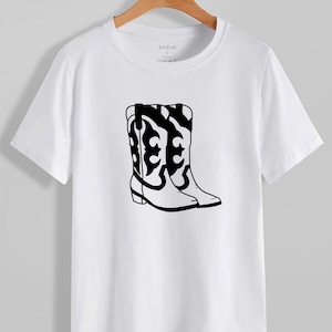 Puede incluir: Camiseta blanca con un gr&aacute;fico negro de botas de vaquero. Las botas tienen dise&ntilde;os decorativos. La marca "SHEIN" es visible en la parte superior. La camiseta est&aacute; colgada en una percha de madera.