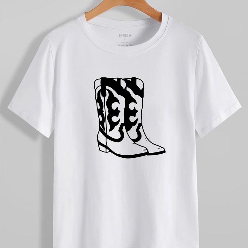 Puede incluir: Camiseta blanca con un gr&aacute;fico negro de botas de vaquero. Las botas tienen dise&ntilde;os decorativos. La marca "SHEIN" es visible en la parte superior. La camiseta est&aacute; colgada en una percha de madera.