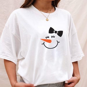 Puede incluir: Camiseta blanca con un dise&ntilde;o de cara de mu&ntilde;eco de nieve. El dise&ntilde;o incluye un lazo negro, pesta&ntilde;as, una nariz de zanahoria naranja y una boca sonriente. La camiseta tiene un corte holgado y de gran tama&ntilde;o.