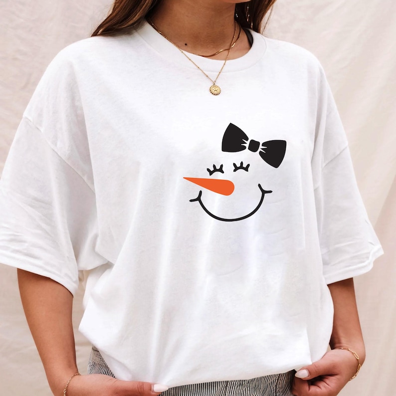 Puede incluir: Camiseta blanca con un dise&ntilde;o de cara de mu&ntilde;eco de nieve. El dise&ntilde;o incluye un lazo negro, pesta&ntilde;as, una nariz de zanahoria naranja y una boca sonriente. La camiseta tiene un corte holgado y de gran tama&ntilde;o.