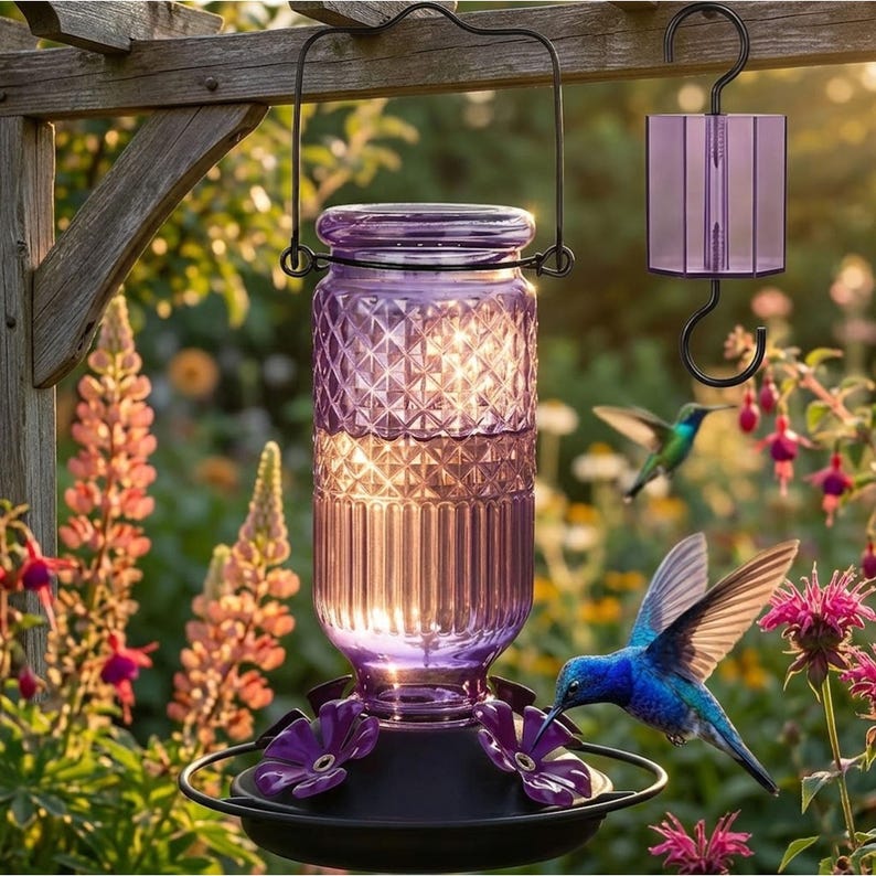 Puede incluir: Un comedero para colibr&iacute;es de vidrio morado con un patr&oacute;n de diamantes en la parte superior y costillas verticales en la inferior. Cuenta con una base de metal negro con puertos de alimentaci&oacute;n en forma de flor y un colgador.