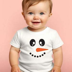 Puede incluir: Una camiseta blanca de manga corta con un dise&ntilde;o de cara de mu&ntilde;eco de nieve. El dise&ntilde;o incluye ojos negros grandes, una nariz de zanahoria naranja y una fila de puntos negros para la boca. La camiseta la lleva un beb&eacute;.