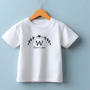 Puede incluir: Camiseta blanca con un dise&ntilde;o gr&aacute;fico negro. El dise&ntilde;o incluye un coraz&oacute;n, hojas y el texto "Richard & Maria" debajo de una "W" estilizada. La camiseta est&aacute; colgada en una percha de madera.