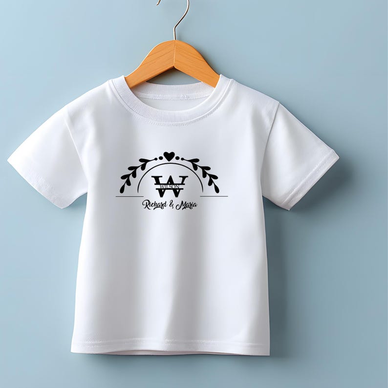 Puede incluir: Camiseta blanca con un dise&ntilde;o gr&aacute;fico negro. El dise&ntilde;o incluye un coraz&oacute;n, hojas y el texto "Richard & Maria" debajo de una "W" estilizada. La camiseta est&aacute; colgada en una percha de madera.