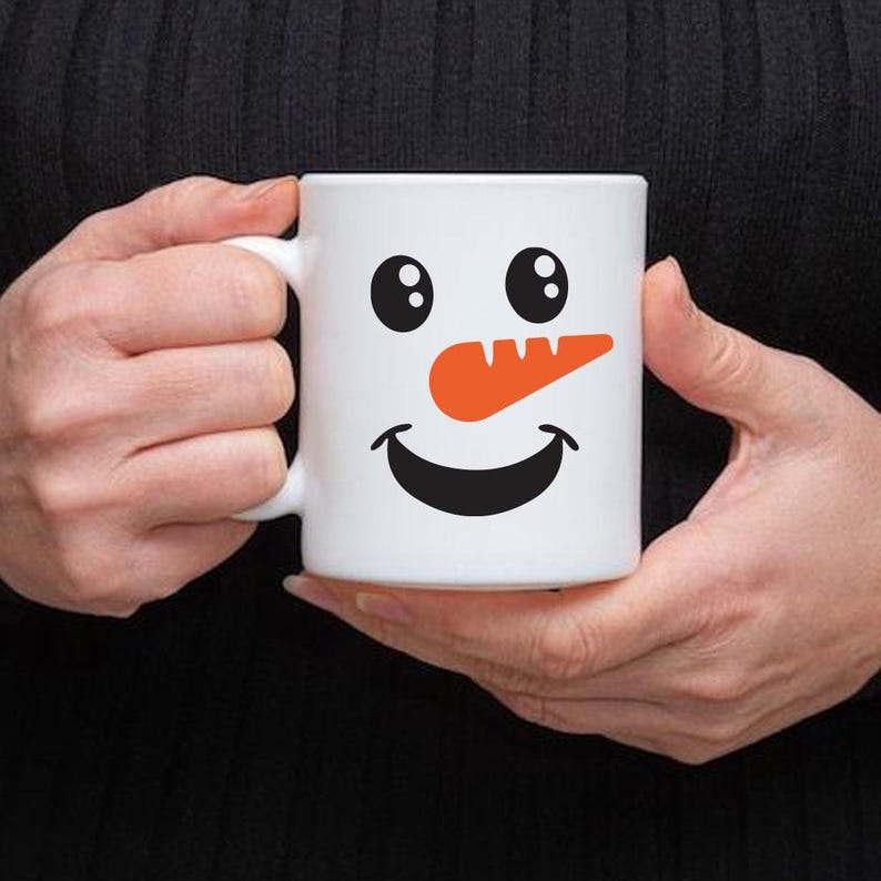 Puede incluir: Taza de cer&aacute;mica blanca con un dise&ntilde;o de cara de mu&ntilde;eco de nieve. La taza tiene ojos negros, una nariz de zanahoria naranja y una boca sonriente negra. La taza es sostenida por una persona.