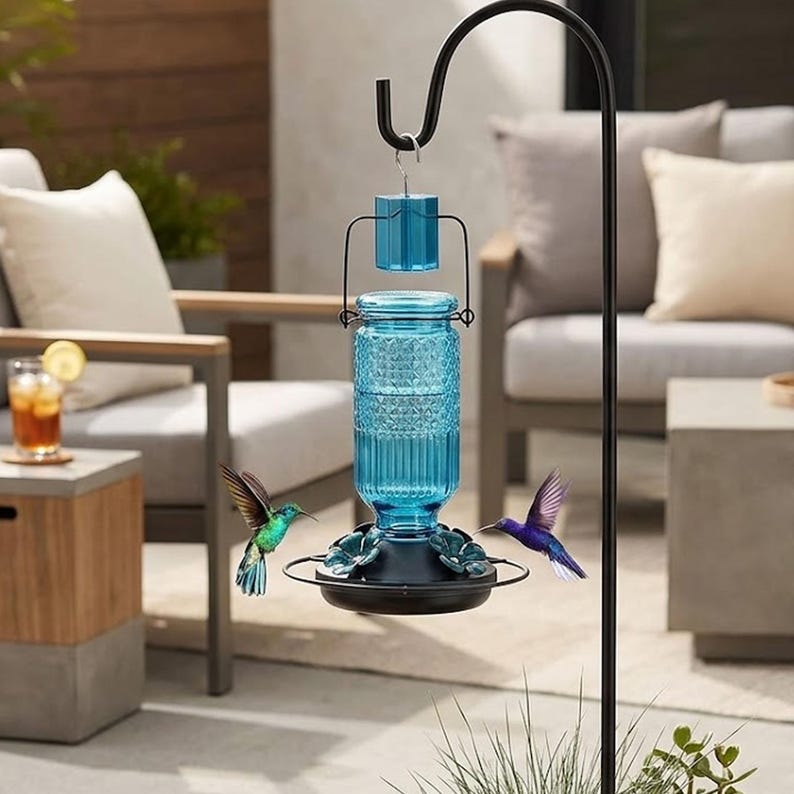 Puede incluir: Un comedero para colibr&iacute;es de cristal azul con un patr&oacute;n de diamantes texturizado, suspendido de un gancho de pastor de metal negro. El comedero tiene una base negra con varios puertos de alimentaci&oacute;n en forma de flor y dos colibr&iacute;es.