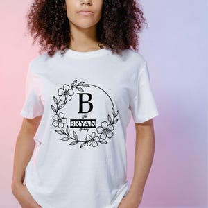 Puede incluir: Camiseta blanca con un dise&ntilde;o de corona floral negra. El dise&ntilde;o incluye la letra "B" y el texto "The Bryan Family". El dise&ntilde;o est&aacute; centrado en la parte delantera de la camiseta.