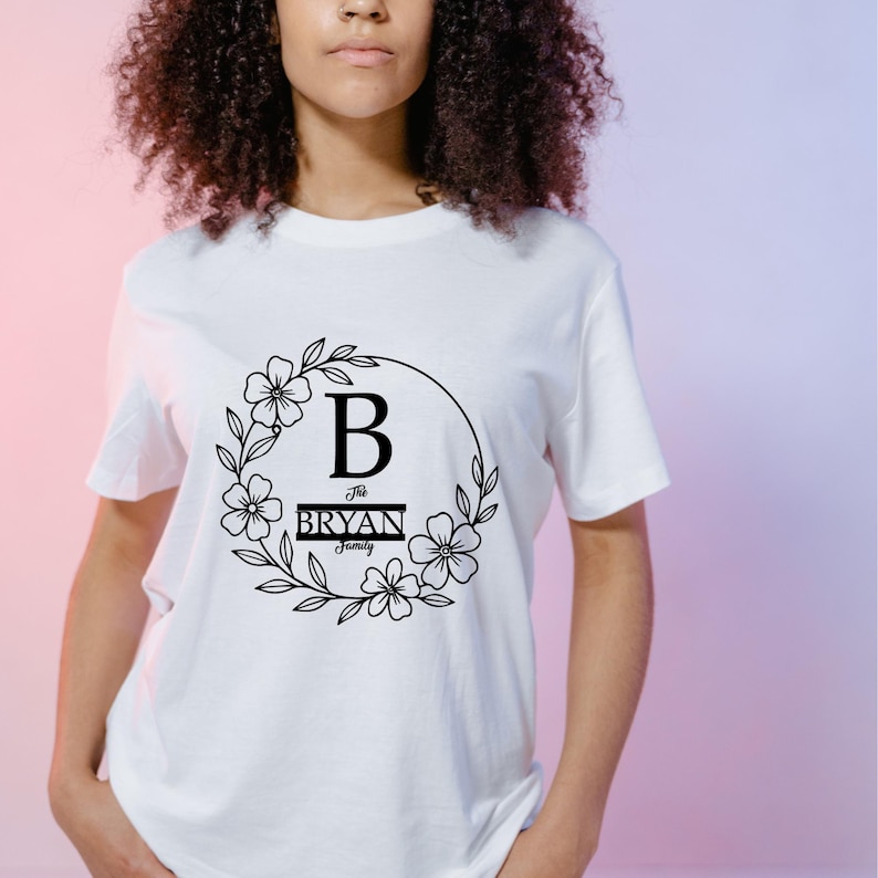 Puede incluir: Camiseta blanca con un dise&ntilde;o de corona floral negra. El dise&ntilde;o incluye la letra "B" y el texto "The Bryan Family". El dise&ntilde;o est&aacute; centrado en la parte delantera de la camiseta.