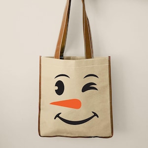 Puede incluir: Un bolso tote beige con la cara de un mu&ntilde;eco de nieve sonriente, con un gui&ntilde;o, cejas negras, una nariz de zanahoria naranja y una boca sonriente. Tiene asas y ribetes de cuero marr&oacute;n.