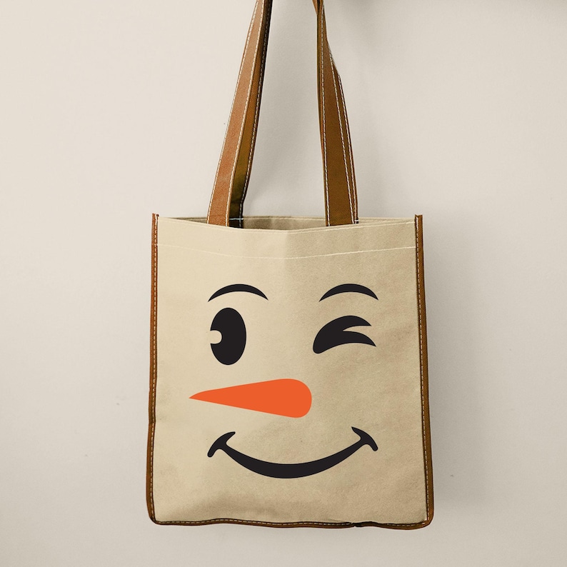Puede incluir: Un bolso tote beige con la cara de un mu&ntilde;eco de nieve sonriente, con un gui&ntilde;o, cejas negras, una nariz de zanahoria naranja y una boca sonriente. Tiene asas y ribetes de cuero marr&oacute;n.
