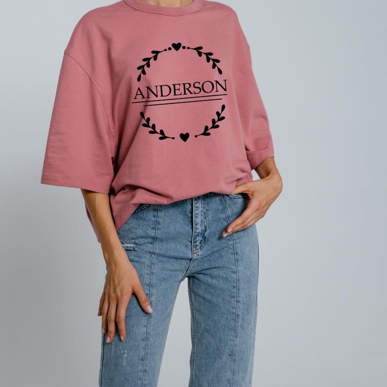 Puede incluir: Camiseta holgada de color rosa empolvado con el nombre "ANDERSON" en negro, enmarcado por una corona decorativa. La camiseta se combina con vaqueros de mezclilla azul claro. El dise&ntilde;o est&aacute; centrado en el pecho.