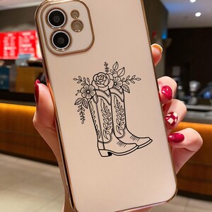 Puede incluir: Una funda de tel&eacute;fono con marco dorado y parte trasera color crema. La parte trasera presenta una ilustraci&oacute;n en blanco y negro de botas de vaquero con detalles florales. La funda tiene recortes para la c&aacute;mara y los botones del tel&eacute;fono.