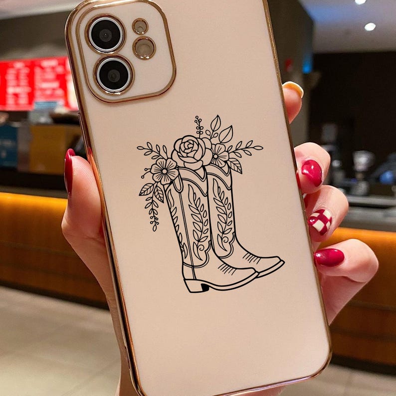 Puede incluir: Una funda de tel&eacute;fono con marco dorado y parte trasera color crema. La parte trasera presenta una ilustraci&oacute;n en blanco y negro de botas de vaquero con detalles florales. La funda tiene recortes para la c&aacute;mara y los botones del tel&eacute;fono.