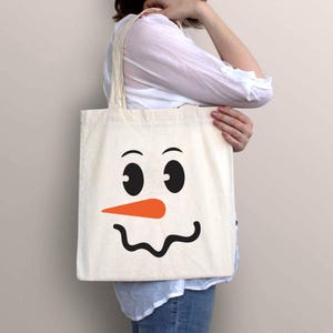 Puede incluir: Una bolsa de mano de color crema con un dise&ntilde;o de cara de mu&ntilde;eco de nieve. La cara del mu&ntilde;eco de nieve tiene ojos negros, una nariz de zanahoria naranja y una boca negra curvada. La bolsa es sostenida por una persona.