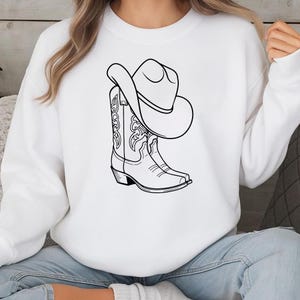 Puede incluir: Sudadera blanca con una ilustraci&oacute;n en blanco y negro de una bota y un sombrero de vaquero. La bota tiene costuras decorativas y el sombrero est&aacute; encima. El dise&ntilde;o es un simple dibujo lineal, perfecto para temas country y western.