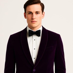 Könnte beinhalten: Ein formeller lila Samtblazer mit schwarzer Fliege und weißem Hemd. Der Blazer hat einen Schalkragen und einen Ein-Knopf-Verschluss. Geeignet für einen besonderen Anlass.