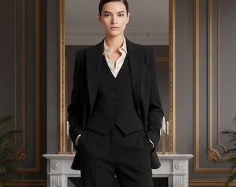 May include: A woman wearing a black three-piece suit, including a blazer, vest, and trousers. The suit is paired with a cream-colored collared shirt. The background features a decorative mirror and a fireplace.