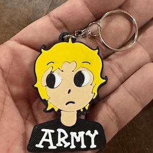 May include: A keychain featuring a cartoon character with bright yellow hair and large black eyes. The character wears a black shirt with the word ARMY printed in white capital letters. The design is made of layered material with a metal ring attached.