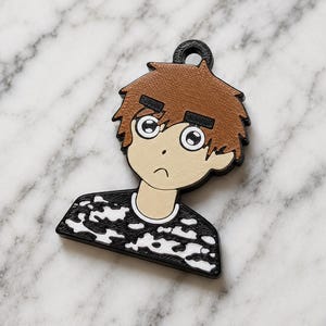May include: A flat, two-dimensional keychain charm featuring a cartoon character with brown hair, large eyes, and a small mouth. The character wears a black shirt with a white camouflage pattern. The charm has a black border and a circular loop at the top.