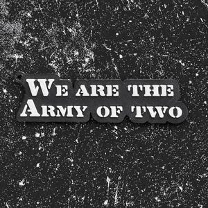 May include: Black and white keychain with the text "We Are The Army Of Two". The text is in a bold, sans-serif font. The keychain is on a textured, black background.