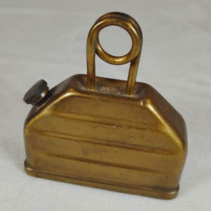Può includere: Un oliatore in metallo color ottone con corpo rettangolare a coste orizzontali, una singola impugnatura circolare sulla parte superiore e un piccolo tappo a vite zigrinato posizionato sul lato superiore sinistro del contenitore.