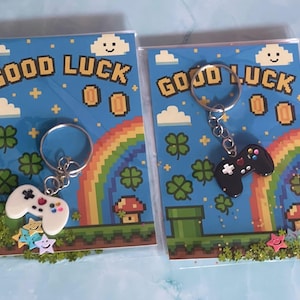Op de afbeelding: Twee sleutelhangers in de vorm van een videogamecontroller, één wit en één zwart, elk met kleurrijke knoppen en een metalen ring. Ze zijn verpakt op rechthoekige kaarten met pixelafbeeldingen van een regenboog, wolken, munten en de tekst GOOD LUCK.