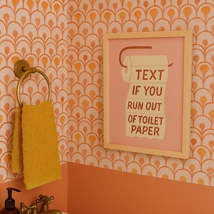 Op de afbeelding: Een badkamer met oranje en perzikkleurige tinten. Een ingelijste print met de tekst "TEXT IF YOU RUN OUT OF TOILET PAPER" hangt boven een kleine witte wastafel met gouden kranen. Een gele handdoek hangt aan een gouden handdoekring.