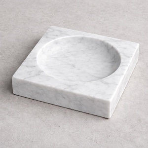 May include: A square, white marble tray with a circular indentation in the center. The marble has gray veining throughout. The tray is a decorative item, suitable for holding small objects.
