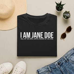 以下が含まれることがあります： 前面に白い文字で「I AM JANE DOE」および「ALL OF US ARE, UNTIL NONE OF US ARE」とプリントされた黒の半袖Tシャツ。麦わら帽子、2つの小さな多肉植物の鉢植え、サングラス、ブルージーンズと一緒に平らに置かれています。