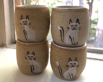 Puede incluir: Cuatro pequeñas tazas de cerámica beige apiladas en un alféizar blanco. Cada taza presenta un diseño de gato blanco pintado a mano con detalles negros. Las tazas tienen un aspecto texturizado y moteado. Fondo: una ventana con árboles.