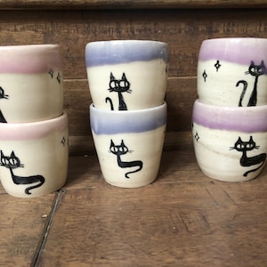 May include: Six ceramic cups with a cream base, each featuring a black cat illustration. The rims are painted in pink, purple, and blue. The cups are stacked in pairs on a wooden surface, with diamond shapes near the cat.