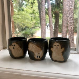 May include: Three black ceramic cups with hand-painted ghost designs. Each cup features a different ghost illustration in shades of beige and white. The cups are displayed on a white windowsill with a blurred outdoor background.