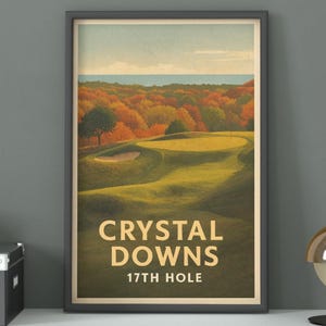 May include: Framed print of a golf course scene with the text "CRYSTAL DOWNS 17TH HOLE". The artwork depicts a green golf course surrounded by trees with autumn foliage. The sky is a light blue, and the frame is dark gray.