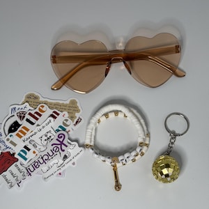 May include: A collection of accessories including heart-shaped sunglasses, a white beaded bracelet with gold accents, a disco ball keychain, and stickers with various phrases and graphics.