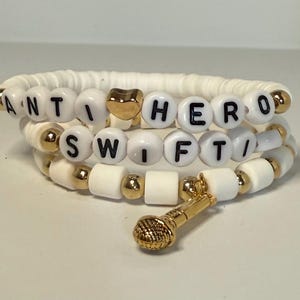 May include: Three-strand bracelet with white beads and gold accents. The bracelet spells out "ANTI HERO SWIFT" with black letter beads. A gold heart bead and a gold microphone charm are also included.
